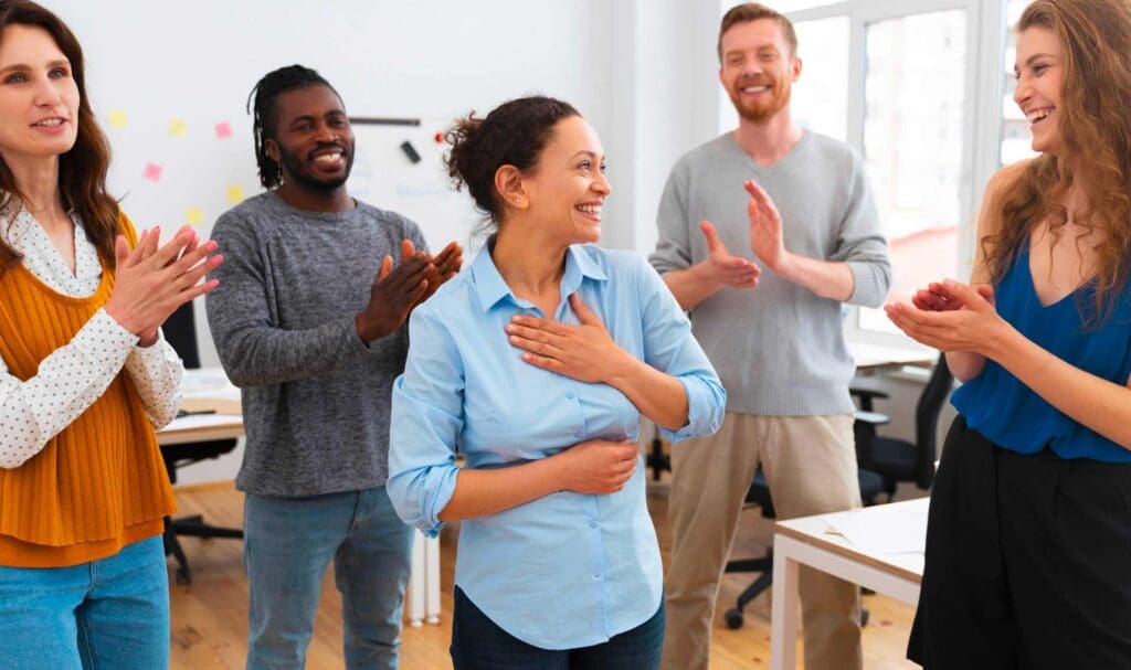 Un groupe de cinq personnes debout dans un bureau, souriant et applaudissant tandis qu'une femme au centre semble heureuse et touchée, la main sur la poitrine.