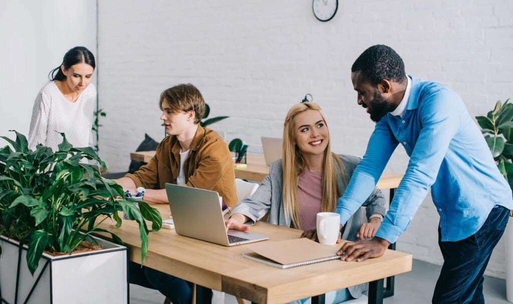 Quatre personnes travaillent ensemble dans un bureau moderne avec des bureaux en bois et des plantes. Deux personnes sont assises à un bureau avec un ordinateur portable, une autre discute debout, et une autre travaille en arrière-plan. L'atmosphère est collaborative et conviviale.