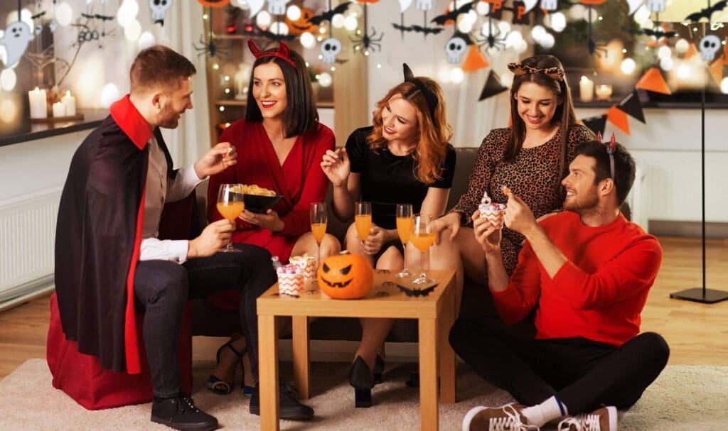 Cinq amis déguisés en Halloween sont assis autour d'une table avec des boissons festives, du pop-corn et une citrouille sculptée. Des décorations d'Halloween, comme des fantômes et des chauves-souris, sont suspendues en arrière-plan. Tout le monde sourit et discute.