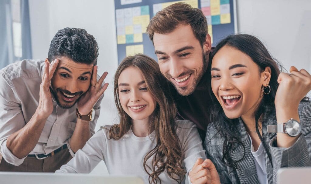 Four young adults gather around a laptop, smiling and cheering with excitement, suggesting they have received good news or achieved a success at work. A colorful sticky note board is visible in the background.
