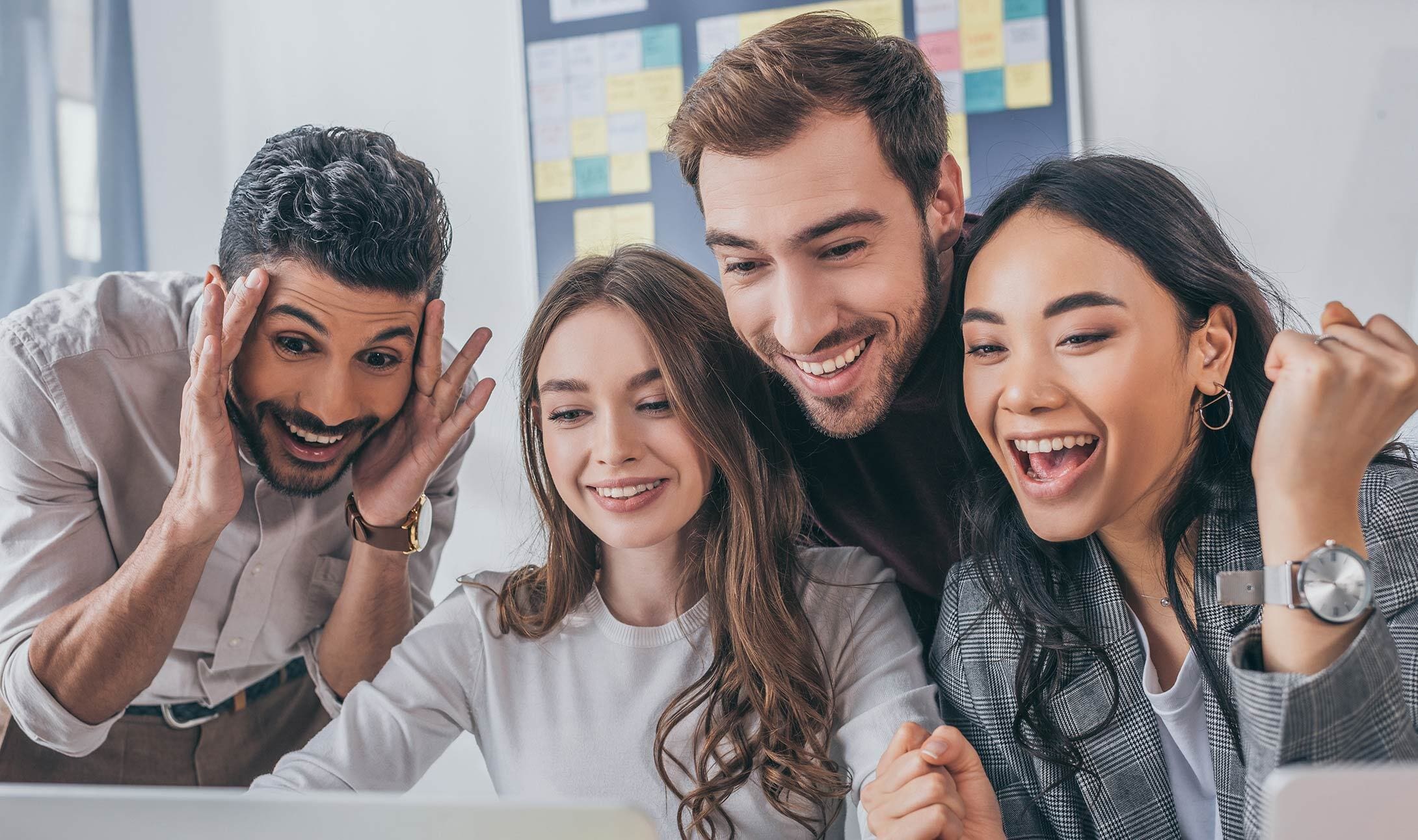 Four young adults gather around a laptop, smiling and cheering with excitement, suggesting they have received good news or achieved a success at work. A colorful sticky note board is visible in the background.