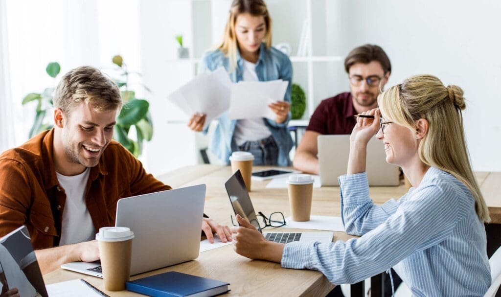 Quatre jeunes adultes travaillent ensemble à une table avec des ordinateurs portables, des papiers et des tasses à café. Deux sont assis et sourient, un est debout en train de lire des documents, et un autre travaille sur un ordinateur portable dans un bureau lumineux.