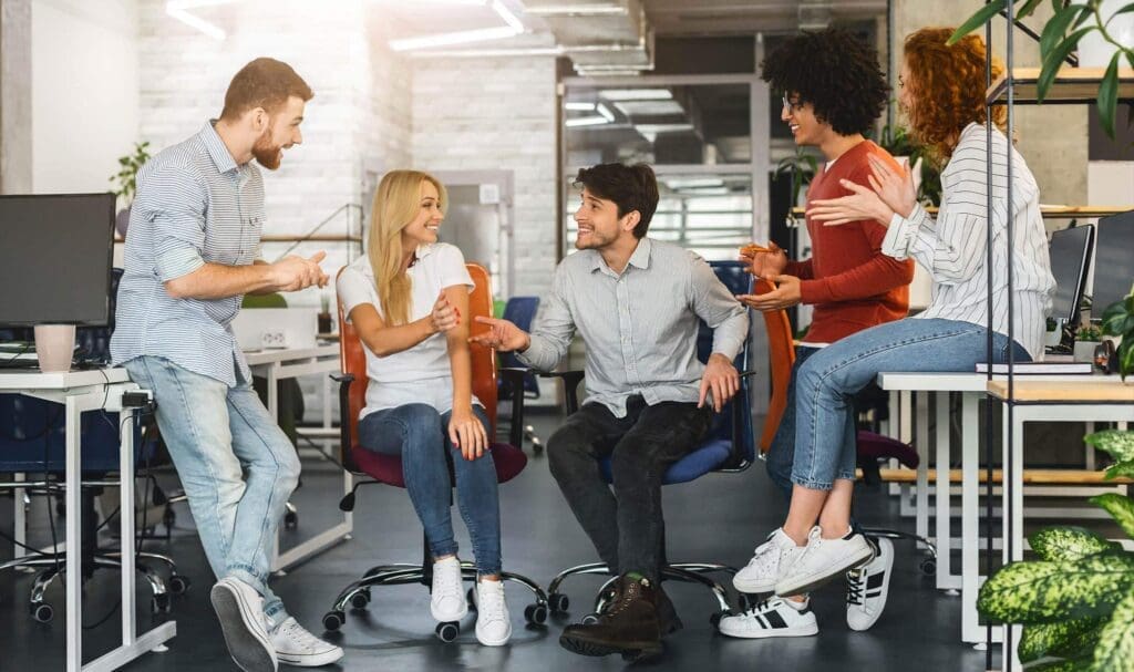 Cinq jeunes adultes en tenue décontractée sourient et discutent ensemble dans un espace de bureau moderne et lumineux, assis sur des chaises et des bureaux, créant une atmosphère conviviale et collaborative.
