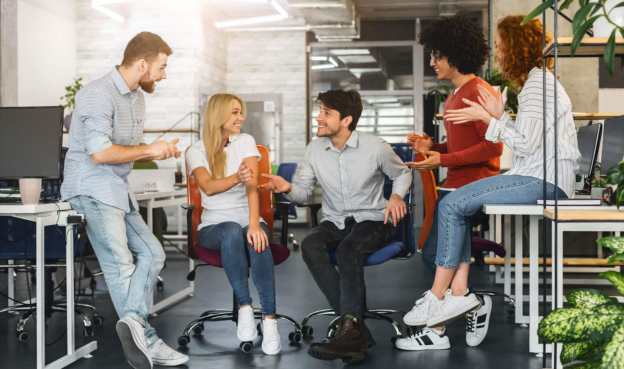 Cinq jeunes adultes en tenue décontractée sourient et discutent ensemble dans un espace de bureau moderne et lumineux, assis sur des chaises et des bureaux, créant une atmosphère conviviale et collaborative.