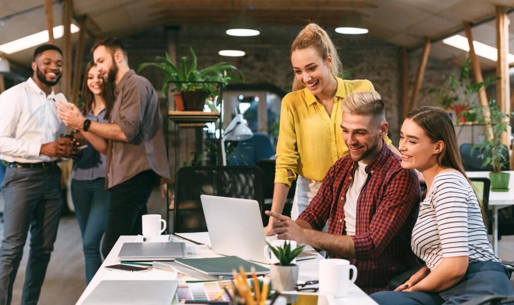 Un groupe de jeunes professionnels dans un bureau moderne sourit tandis que trois personnes collaborent autour d'un ordinateur portable, tandis que trois autres interagissent et rient en arrière-plan. L'espace de travail est lumineux et décoré de plantes.