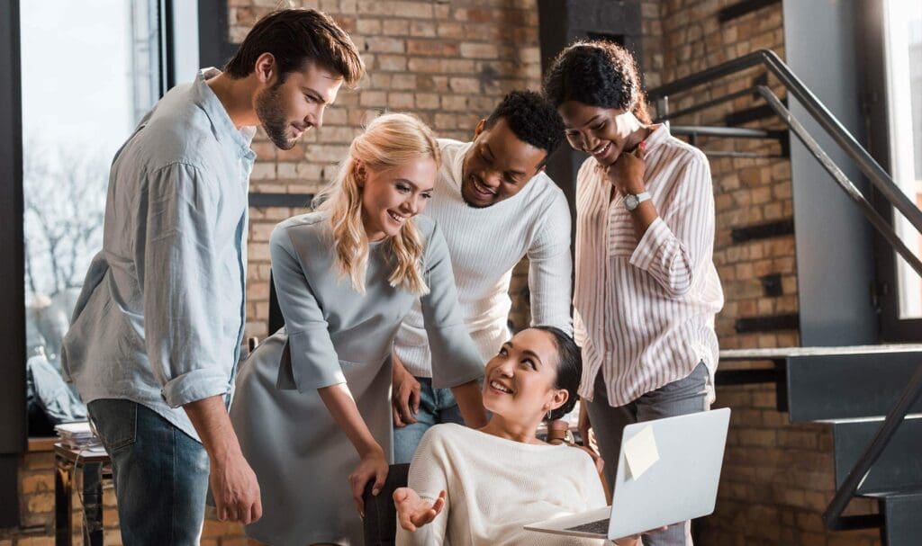 Cinq personnes se rassemblent autour d'un ordinateur portable, souriantes et engagées dans une conversation, dans un bureau moderne avec des murs en briques et de grandes fenêtres.