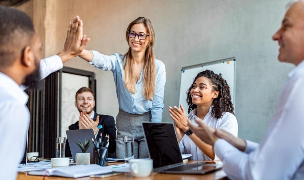 Une femme souriante debout et donnant un high-five à un collègue dans un bureau lumineux, tandis que trois collègues sont assis autour d'une table, applaudissant et souriant pendant une réunion.