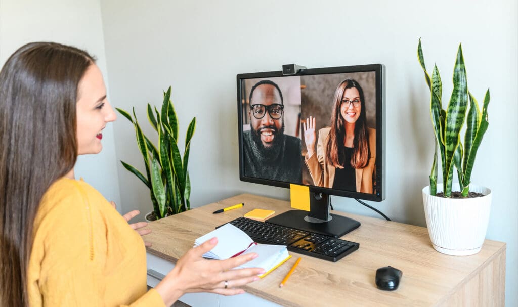 A woman sits at a desk, smiling and gesturing while video chatting with a man and a woman on her computer screen. The desk has notebooks, pens, a keyboard, and is decorated with potted plants.