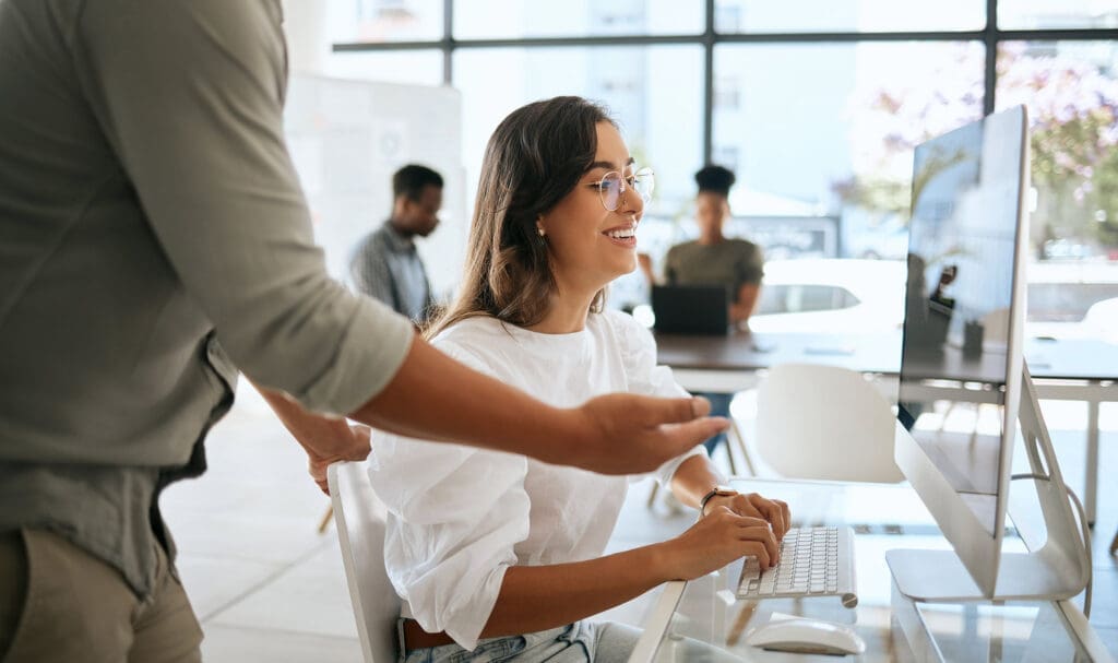 Une femme portant des lunettes sourit en tapant sur un clavier à son bureau, tandis qu'un collègue se tient à côté d'elle et lui fait des gestes. Deux autres personnes travaillent en arrière-plan dans un bureau moderne et lumineux.
