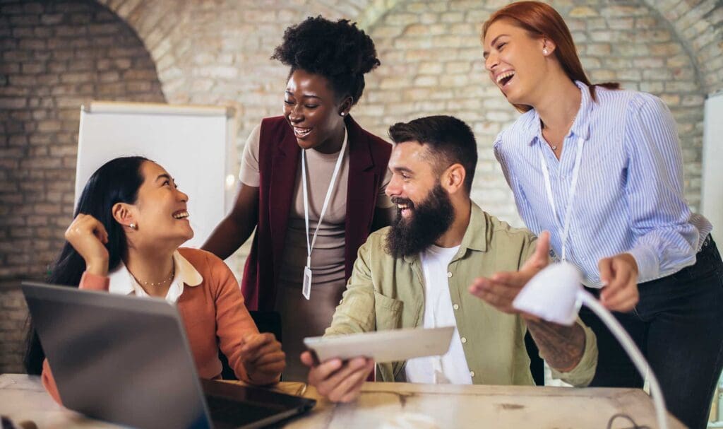 Four coworkers gather around a desk, smiling and laughing together. One person holds a tablet, while another sits in front of a laptop. They appear to be enjoying a lively team discussion in a modern office.