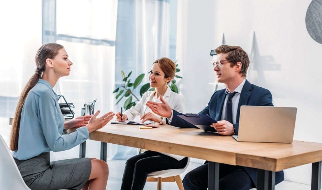 Trois personnes en tenue professionnelle sont assises à un bureau dans un bureau lumineux, une femme étant interviewée par un homme et une femme qui prennent des notes et tiennent un presse-papiers et un ordinateur portable.