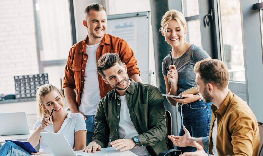 Five young adults sit and stand around a table, smiling and talking in a bright office setting with a laptop and documents in front of them, suggesting a collaborative work meeting.