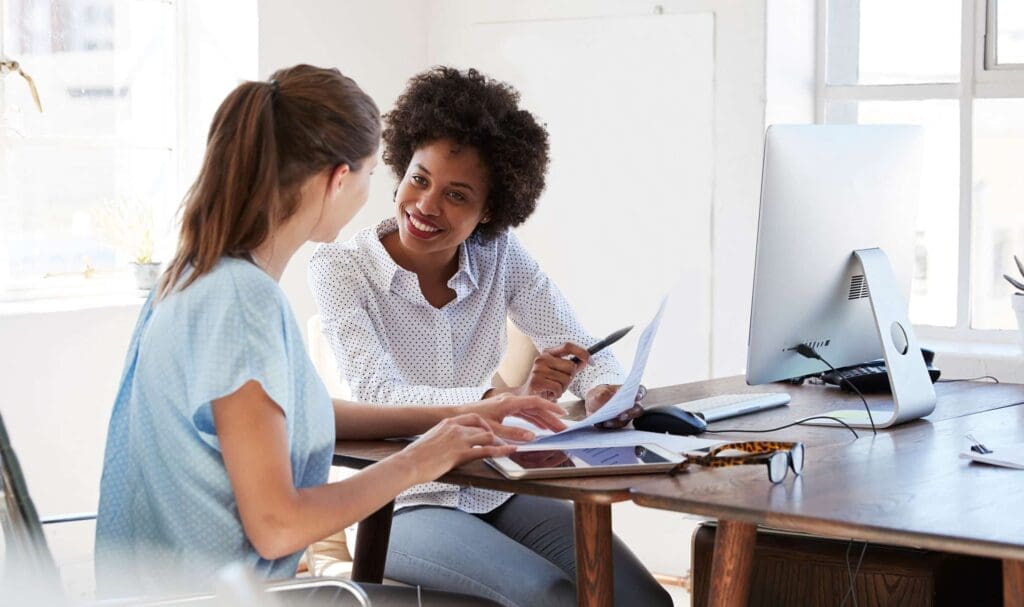 Deux femmes sont assises à un bureau lumineux, souriantes et discutant. L'une pointe un document tandis que l'autre tape sur un ordinateur portable. Des papiers, un écran d'ordinateur et des lunettes sont posés sur le bureau.