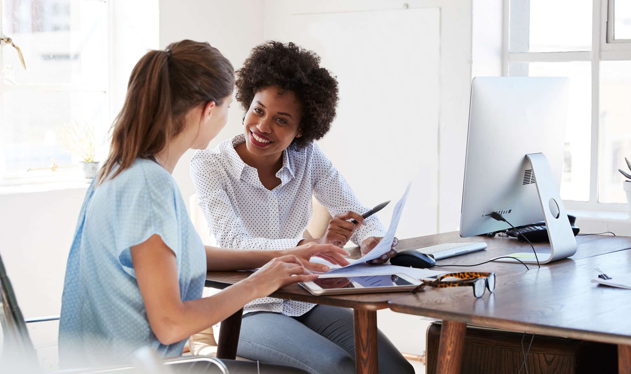 Deux femmes sont assises à un bureau lumineux, souriantes et discutant. L'une pointe un document tandis que l'autre tape sur un ordinateur portable. Des papiers, un écran d'ordinateur et des lunettes sont posés sur le bureau.