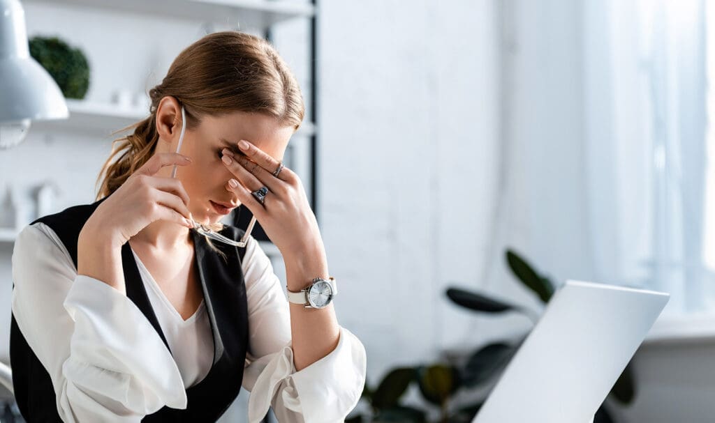 Une femme assise à un bureau tient ses lunettes et se pince l'arête du nez, semblant stressée ou fatiguée, avec un ordinateur portable devant elle dans un environnement de bureau lumineux.