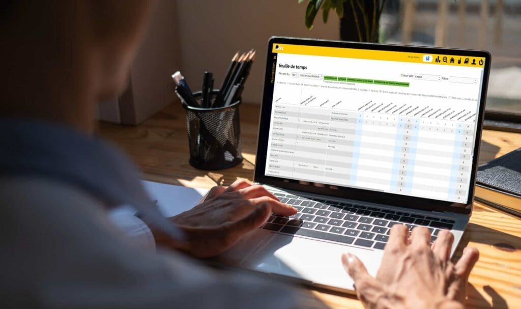 Une personne utilisant un ordinateur portable à un bureau, consultant une feuille de temps ou une application de suivi des heures de travail avec un tableau et diverses colonnes, y compris des dates et des valeurs numériques.