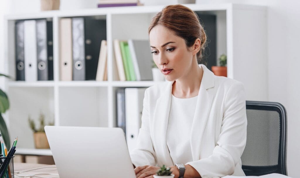 Une femme en blazer blanc est assise à un bureau, concentrée sur son ordinateur portable. Derrière elle, des étagères remplies de classeurs et de fournitures de bureau créent un environnement de travail professionnel.