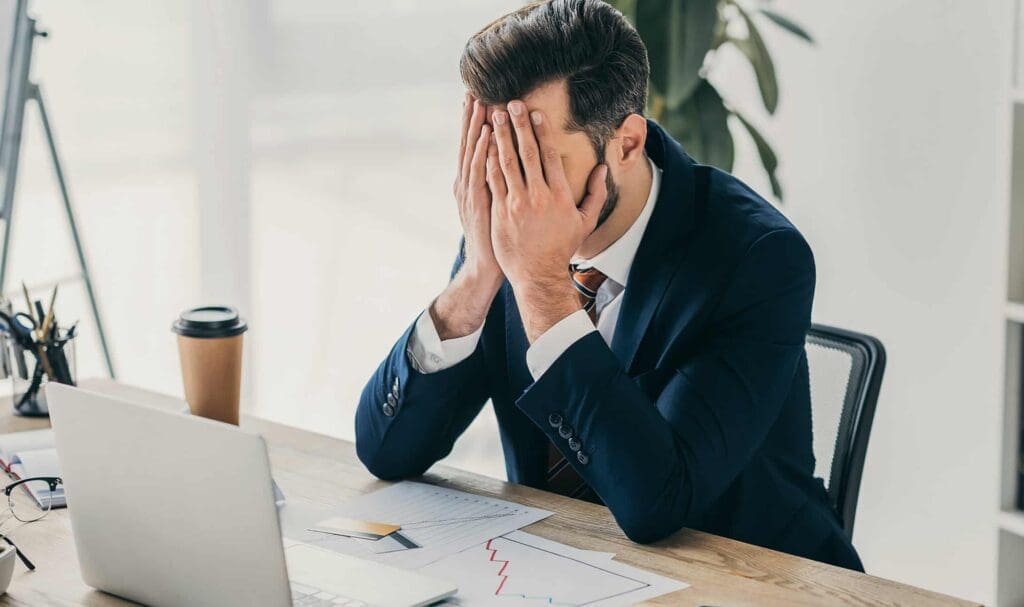 Un homme en costume est assis à un bureau, les mains sur le visage, l'air stressé. Un ordinateur portable ouvert, des documents avec des graphiques, un stylo, un carnet et une tasse de café à emporter sont posés sur le bureau.
