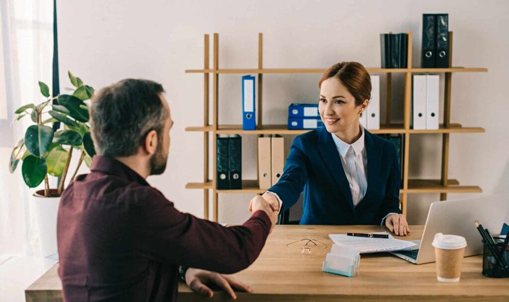 Une femme en tailleur est assise à un bureau, souriante et serrant la main d'un homme en face d'elle. Des étagères, des dossiers, un ordinateur portable et une tasse à café sont visibles en arrière-plan.