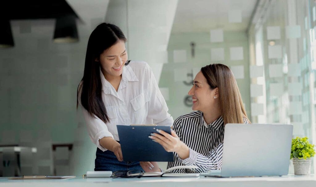 Deux femmes dans un bureau moderne sourient et discutent de documents. L'une est assise à un bureau avec un ordinateur portable, tandis que l'autre se tient à côté d'elle, un presse-papiers à la main. La lumière naturelle baigne la pièce grâce à de grandes fenêtres.