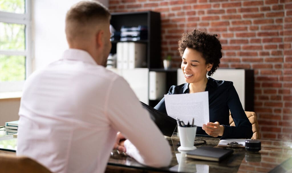 Une femme sourit tout en tenant un document et en parlant à un homme à travers un bureau dans un bureau moderne avec des murs en briques apparentes et des étagères en arrière-plan.