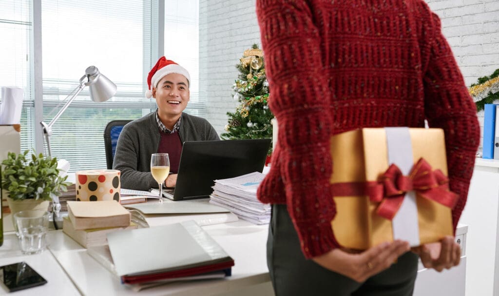Une personne portant un pull rouge tient un cadeau emballé derrière son dos, s'approchant d'un homme souriant portant un chapeau de Père Noël à son bureau, avec un arbre de Noël en arrière-plan.