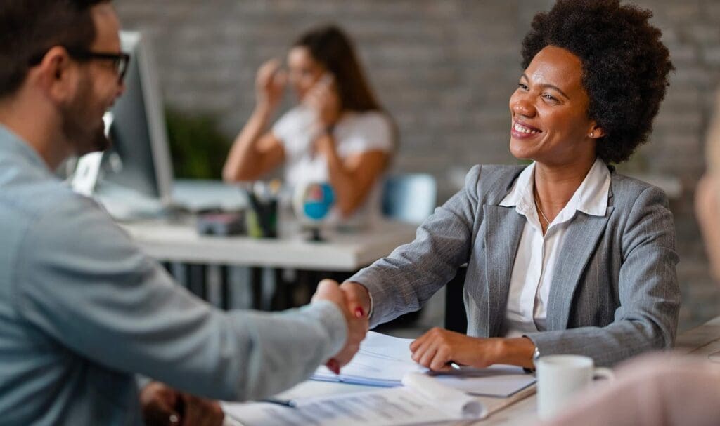 Une femme souriante, vêtue d'un blazer gris, serre la main d'un homme assis à un bureau, avec des papiers et une tasse de café devant elle. D'autres personnes travaillent en arrière-plan.