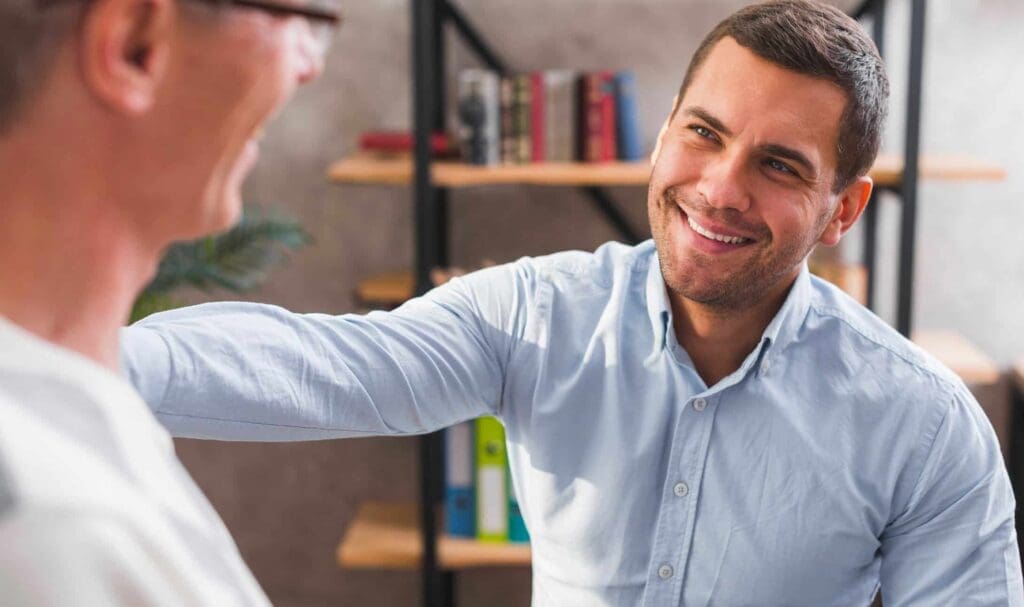 Un homme souriant, vêtu d'une chemise bleu clair, est assis à l'intérieur et pose doucement sa main sur l'épaule d'un autre homme, lui offrant soutien et encouragement. Des étagères et des livres sont visibles en arrière-plan.
