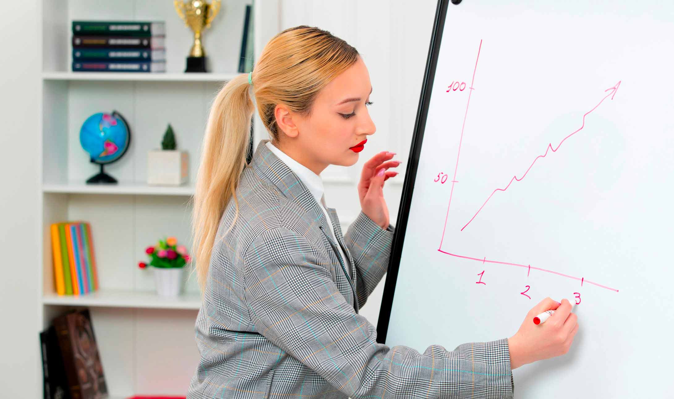 Dans un bureau, une femme en blazer gris dessine un graphique linéaire à la hausse sur un tableau blanc, indiquant une croissance. Des livres, un trophée et un globe terrestre sont visibles sur des étagères en arrière-plan.