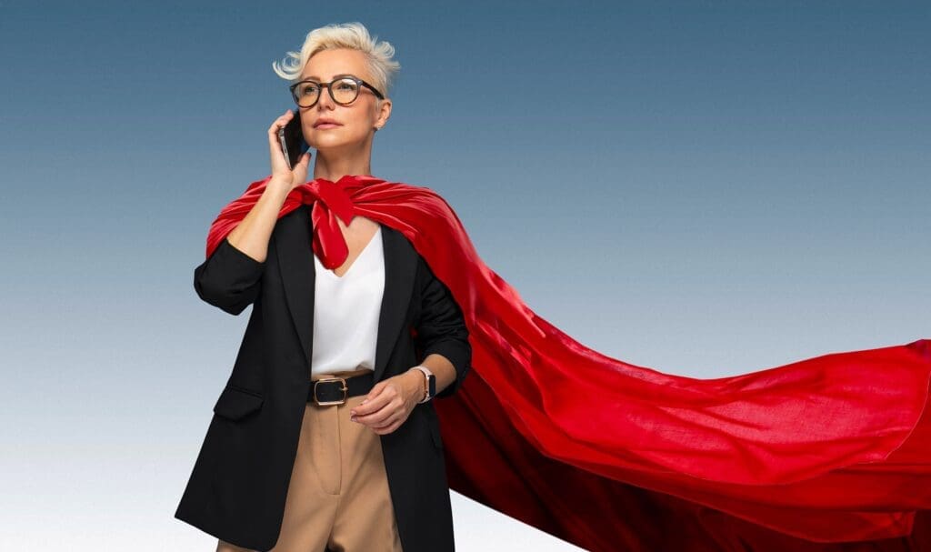 Une femme confiante aux cheveux blonds courts portant des lunettes, un costume et une cape rouge fluide, se tient debout en train de parler sur un smartphone sur un fond bleu dégradé.