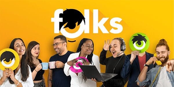 Un groupe diversifié de personnes souriantes, tenant des accessoires colorés, se tient devant un fond jaune vif où le mot « folks » est écrit en lettres blanches et grasses. Certains tiennent des symboles, l'un utilise un ordinateur portable et un autre parle sur un téléphone jaune.