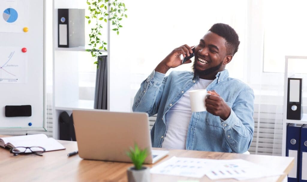 Un homme est assis à un bureau avec un ordinateur portable, souriant tout en parlant au téléphone et tenant une tasse blanche. L'espace de travail est lumineux, avec des papiers, une plante et des fournitures de bureau bien visibles.