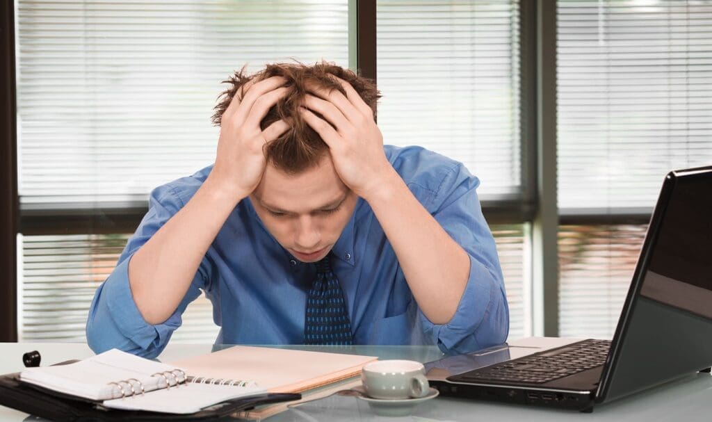 Un homme stressé en chemise bleue est assis à un bureau, la tête dans les mains, entouré d'un ordinateur portable ouvert, d'un cahier, de documents et d'une tasse, dans un environnement de bureau lumineux.