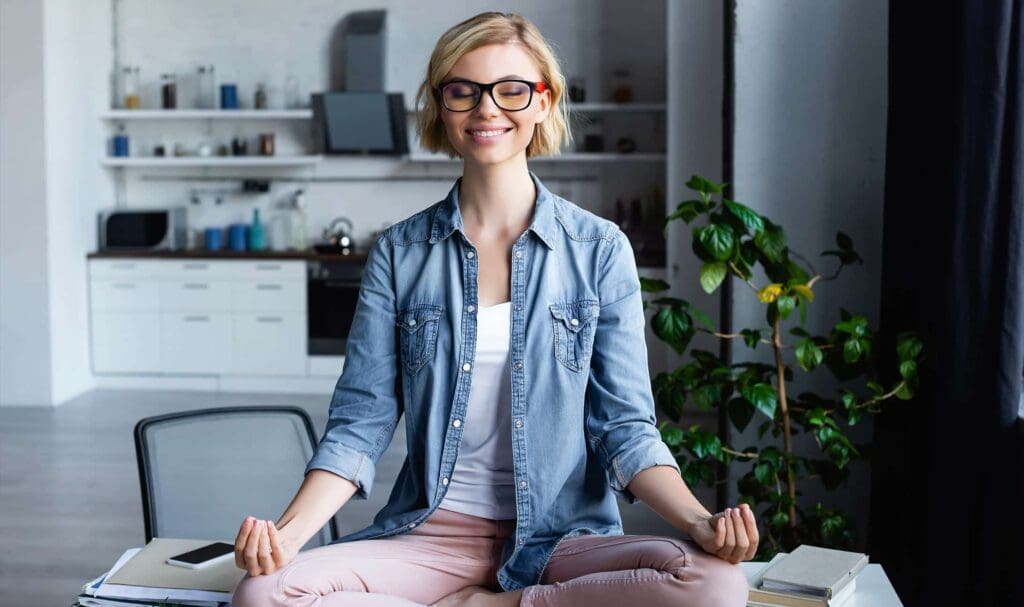 A smiling woman with glasses sits cross-legged on a desk in a modern kitchen, meditating with her eyes closed and hands resting on her knees, surrounded by books and a potted plant.