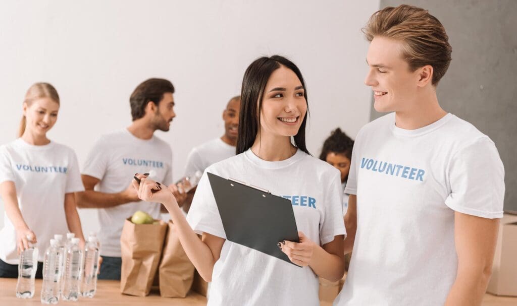 Un groupe de jeunes adultes portant des t-shirts « bénévoles » travaillent ensemble, souriants. Une femme tient un presse-papiers et un stylo et discute avec un homme, tandis que d'autres trient des provisions et des bouteilles d'eau en arrière-plan.