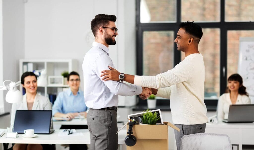 Deux hommes se serrent la main et sourient dans un bureau, l'un tenant un carton rempli d'affaires, suggérant un premier jour de travail. Trois collègues assis à leurs bureaux en arrière-plan, observent et sourient.