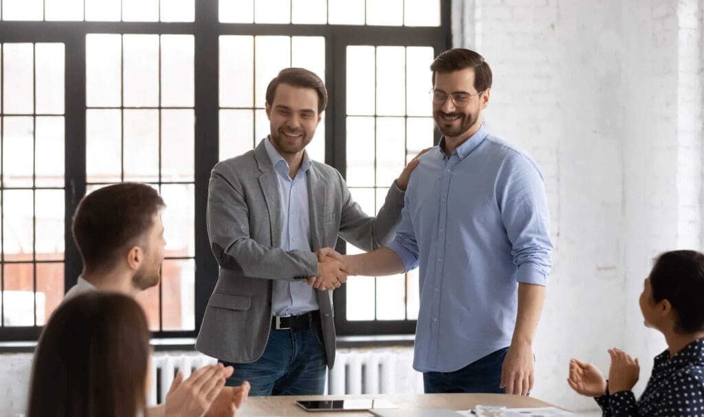 Deux hommes se tiennent debout et se serrent la main en souriant, tandis que trois collègues assis à une table les applaudissent dans un bureau lumineux avec de grandes fenêtres en arrière-plan.