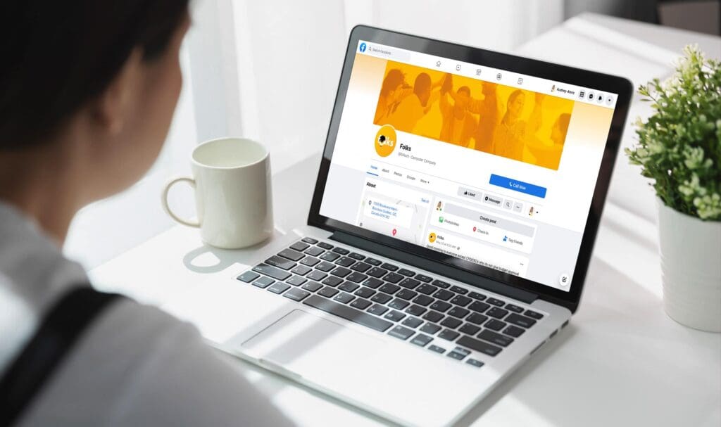 Une personne assise à un bureau, une tasse blanche et une plante à la main, consulte son profil sur un réseau social sur un ordinateur portable. L'écran affiche une page avec une bannière orange et une photo de profil.