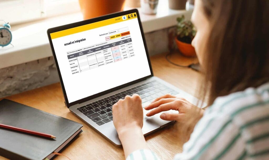 Une personne utilise un ordinateur portable affichant une page de gestion d'inscription de compte avec un tableau des informations utilisateur et des statuts. Elle est assise à un bureau en bois près d'une fenêtre, un carnet et un stylo à côté d'elle.
