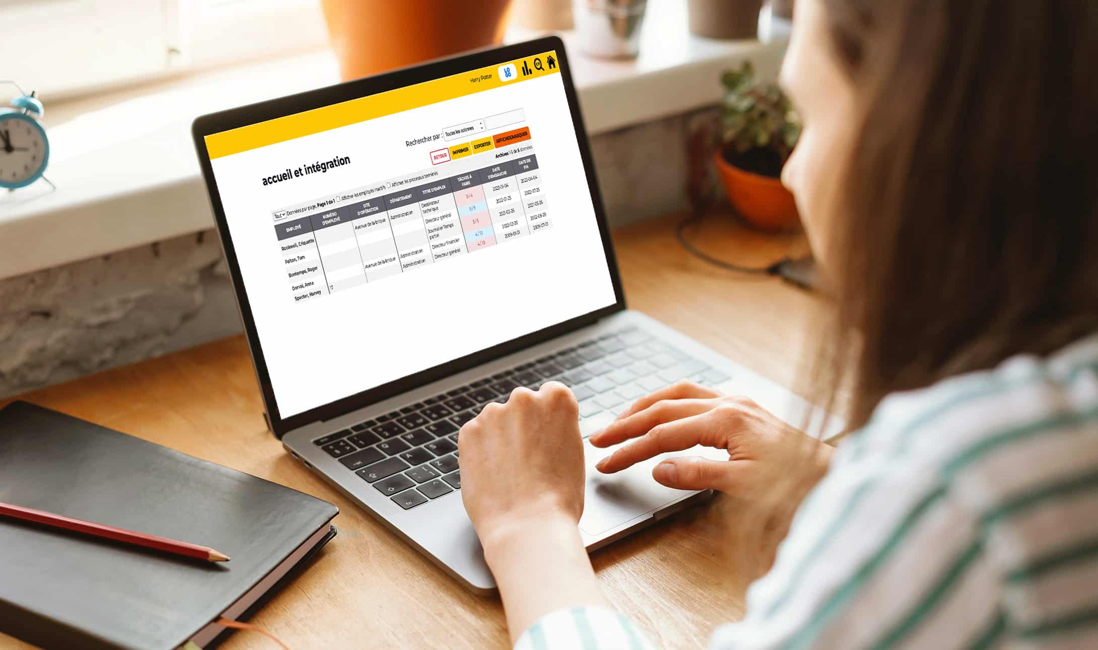Une personne utilise un ordinateur portable affichant une page de gestion d'inscription de compte avec un tableau des informations utilisateur et des statuts. Elle est assise à un bureau en bois près d'une fenêtre, un carnet et un stylo à côté d'elle.
