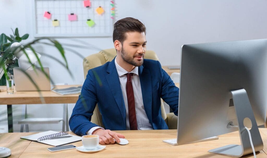 Un homme en costume bleu marine et cravate est assis à un bureau dans un bureau moderne, travaillant sur ordinateur. Un carnet, un stylo et une tasse de café sont posés sur le bureau. Un tableau blanc avec des post-its est visible en arrière-plan.