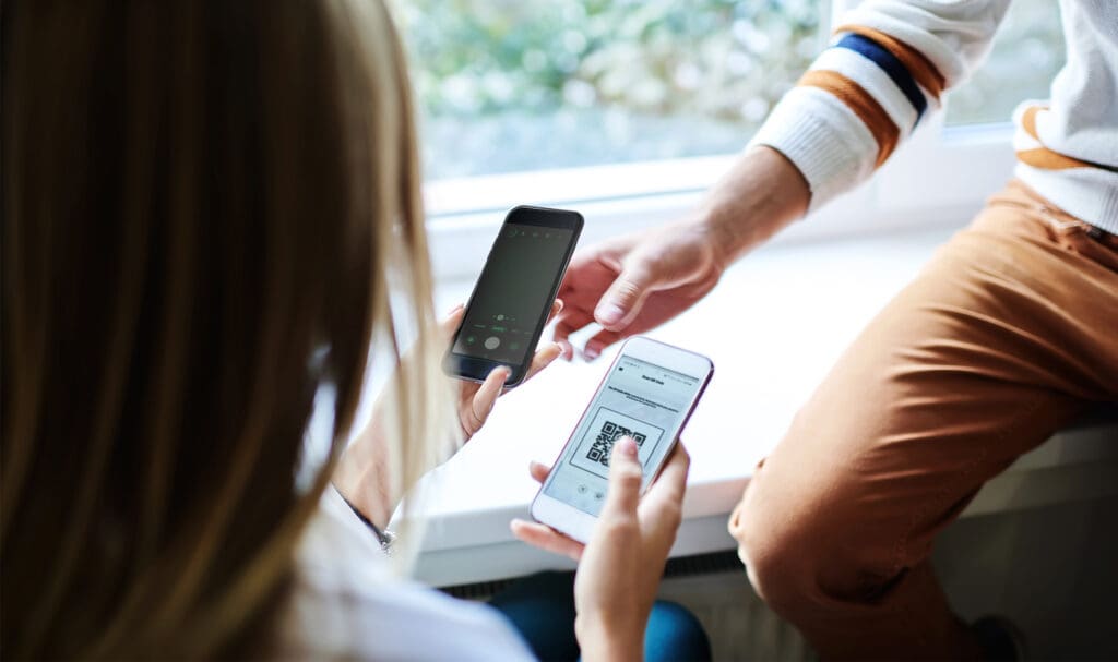 Two people are sitting near a window. One is holding a phone displaying a QR code, while the other is holding a phone, possibly about to scan the code. Both are dressed casually.