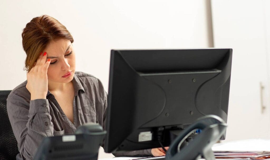 Une femme est assise à un bureau devant un ordinateur, la tête appuyée sur sa main, l'air stressée ou fatiguée. Des papiers et un téléphone sont également visibles sur le bureau.