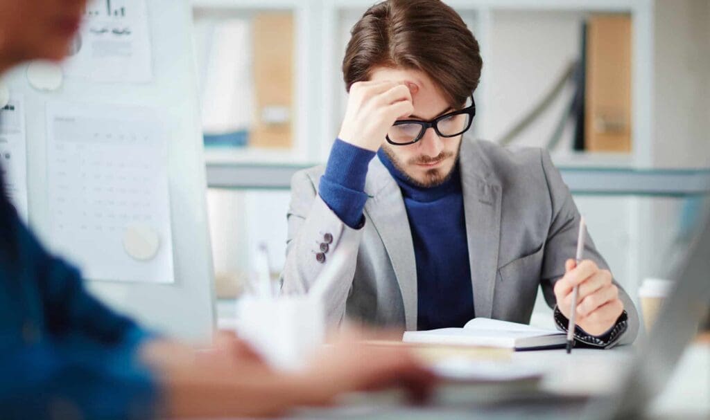 Un homme portant des lunettes et un blazer est assis à un bureau, le front baissé, concentré, tout en lisant et en prenant des notes. Des documents et des collègues sont visibles en arrière-plan.