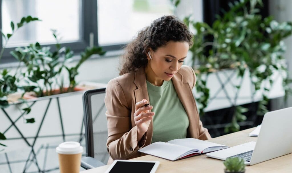 Une femme aux cheveux bouclés, vêtue d'un blazer marron, est assise à un bureau avec un carnet, un ordinateur portable et une tablette. Elle écrit tout en écoutant avec des écouteurs sans fil. Des plantes et une tasse de café sont posées sur le bureau.