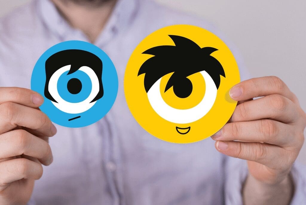 Une personne tient deux autocollants ronds représentant des visages de dessin animé : l'un bleu avec une expression triste, l'autre jaune avec une expression joyeuse. Les autocollants représentent de grands yeux simples et des cheveux stylisés.