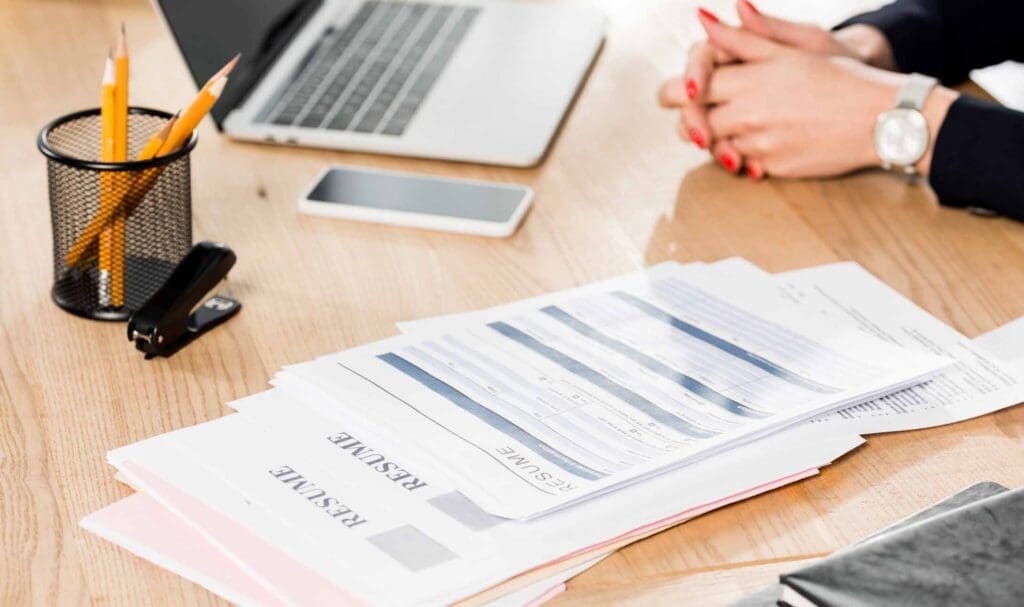 Un bureau avec une pile de CV, un ordinateur portable, un smartphone, des crayons dans un support, une agrafeuse et une personne avec les mains jointes et du vernis à ongles rouge assise à la table.