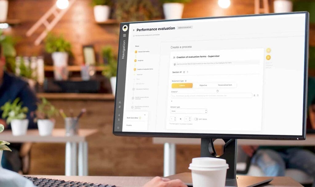 Une personne travaille à un bureau avec une tasse de café, regardant un écran d'ordinateur affichant une interface de formulaire d'évaluation des performances dans un bureau avec des collègues flous et des plantes en arrière-plan.