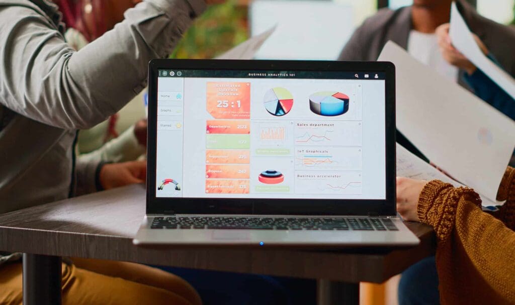 A laptop on a table displays colorful business analytics charts and graphs while three people sit around, holding papers and discussing.