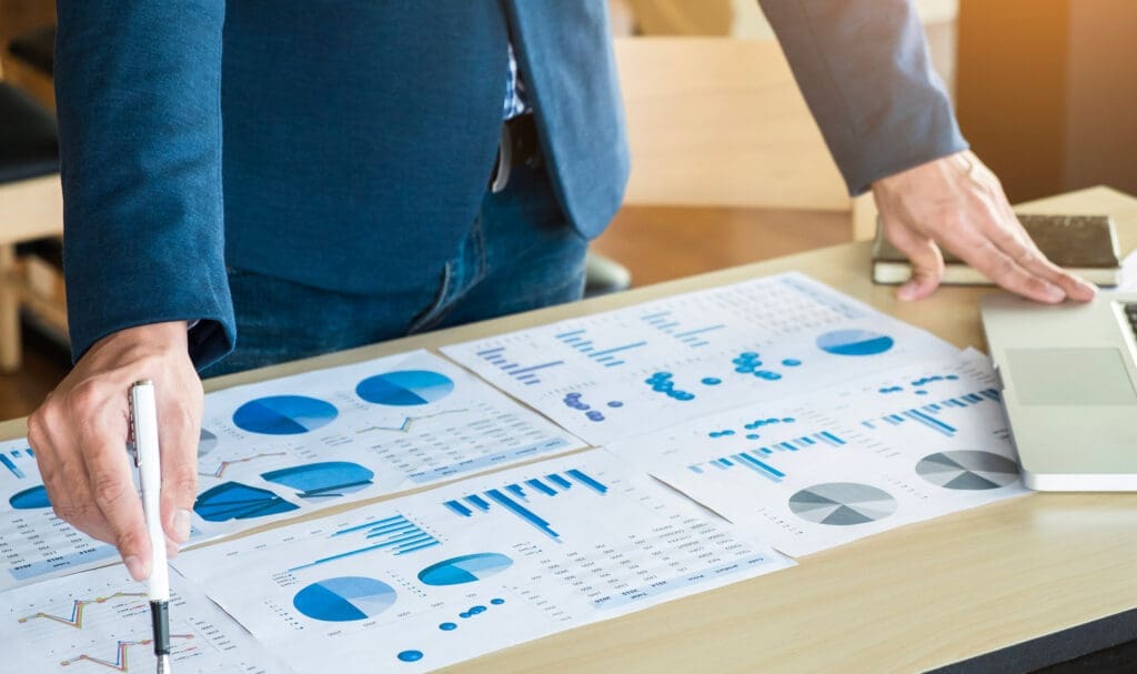 Une personne en costume se tient à un bureau, examinant et pointant du doigt des graphiques et des diagrammes imprimés aux éléments bleus, étalés sur la table. Un ordinateur portable et un smartphone sont également visibles.